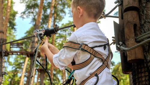 Closeup of little boy clamping safety rope with hook before riding on zip line. Active childhood, healthy lifestyle, kids playing outdoors, children in nature.
