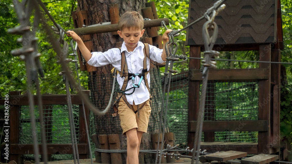 Obraz premium Cute boy having fun while walking on wobbly rope bridge at adventure park. Active childhood, healthy lifestyle, kids playing outdoors, children in nature.