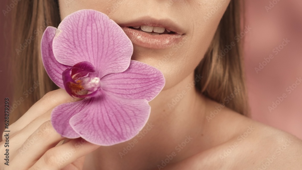 Female lips with delicate pink lipstick and orchid flower closeup on a pink blurred background. A beautiful smile. Perfect female face makeup. Lipstick. The concept of beauty, cosmetology, skin care. Female lips with delicate pink lipstick and orchid flower closeup on a pink blurred background. A beautiful smile. Perfect female face makeup. Lipstick. The concept of beauty, cosmetology, skin care.