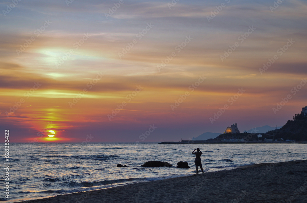 Silhouette of a woman standing near the sea, looking on the sunset and making foto. Amazing sunset at the sea with view on a castle