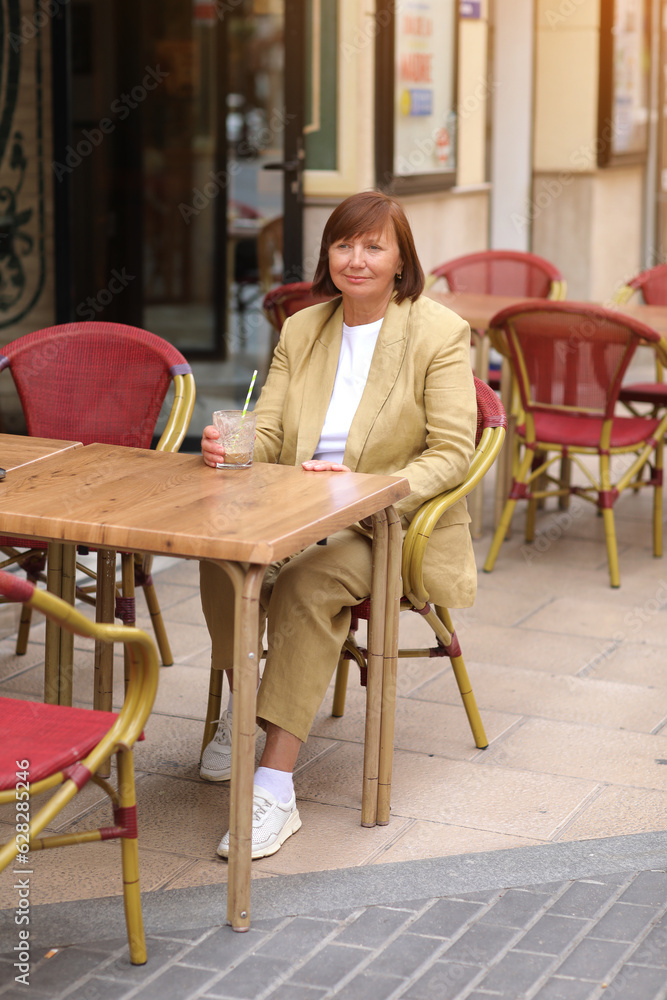 Attractive stylish mature woman 50-60 s with short hair in yellow jacket is drinking coffee milk drink with straw and relaxing at cafe in European city street. Concept of summer holiday vacation