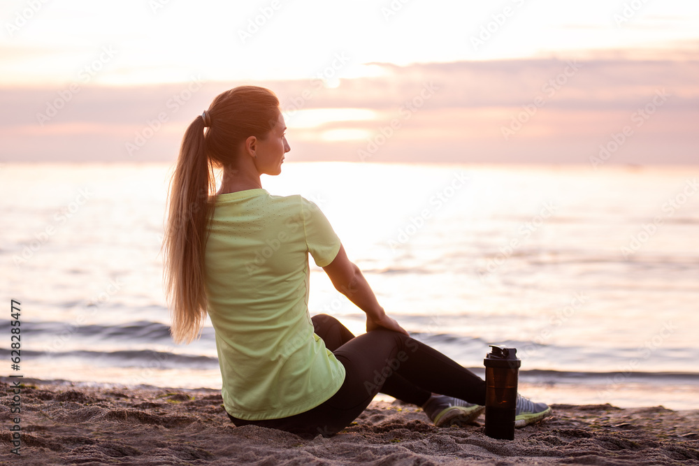 Young woman in sportswear is sitting on the seashore at sunrise. Girl is resting after workout Young woman in sportswear is sitting on the seashore at sunrise. Girl is resting after workout