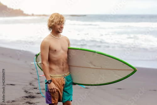 Fit young surfer man with curly blond hair with surfboard goes by the ocean having fun doing extreme water sports, surfing. Travel and healthy lifestyle concept. Sports travel destination.