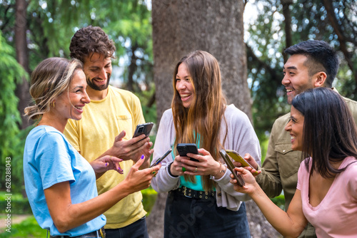 Smiling multiethnic group with phones watching internet or social networks in the park, technology concept