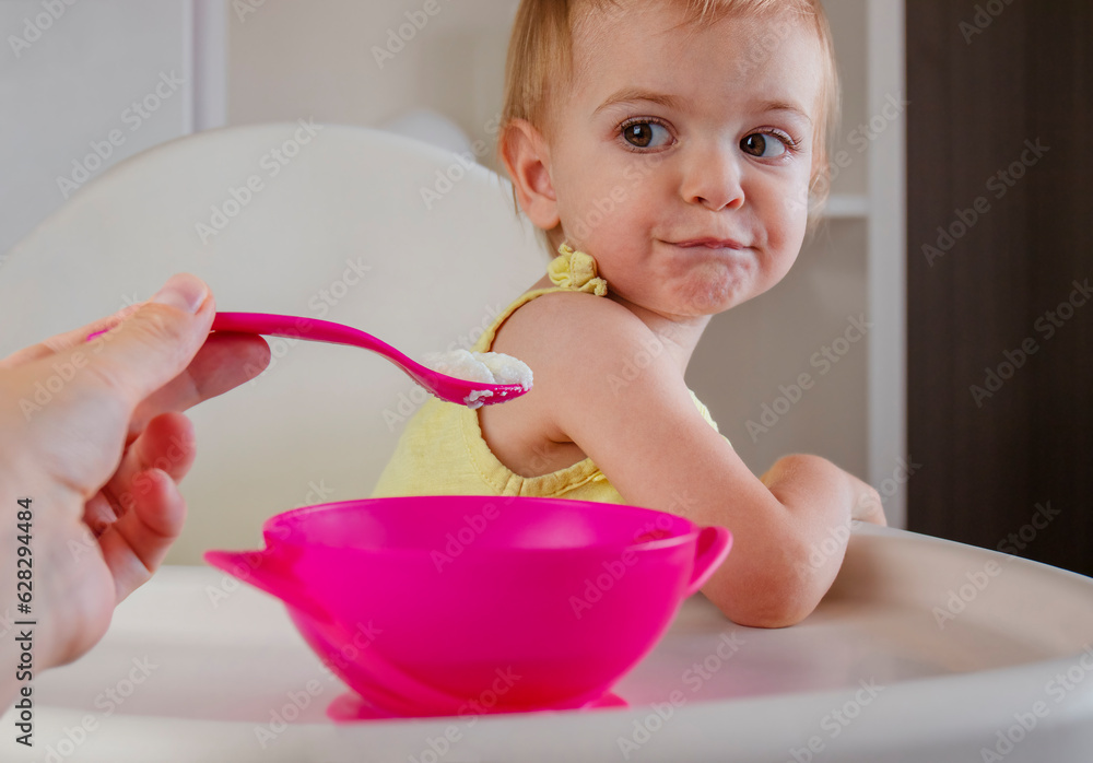 Toddler girl sitting in high chair rejecting eating porridge. Baby Feeding. Mother feeding child Toddler girl sitting in high chair rejecting eating porridge. Baby Feeding. Mother feeding child