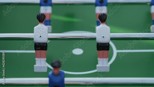 Figures of football players on metal axes rotate in different directions and hit game ball in kicker. Game of table football by blue and white team closeup in natural light in center of green pitch. photo