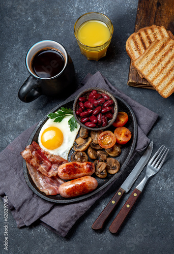 Full fry up English breakfast with fried eggs, sausages, bacon, black pudding, beans, toast and coffee, dark background