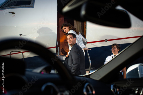 Successful couple boarding private Jet , man looking back over shoulder on tarmac of airport, view from drivers seat of sports car parked next to plane on tarmac  photo