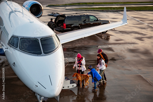High angle view of Family with dog boarding private plane on wet tarmac of private airport for family vacation , pilot in background unloading luggage from SUV 