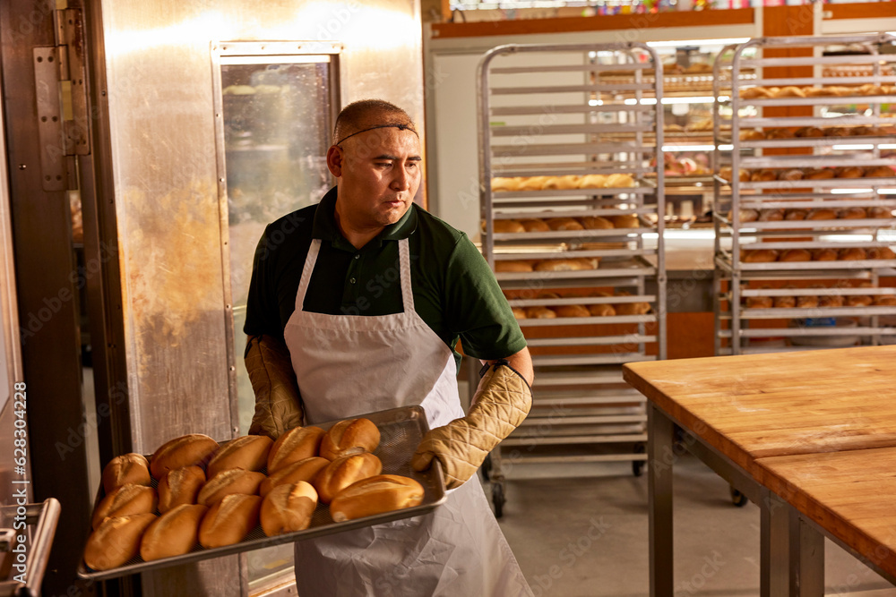 Hispanic baker in a commercial kitchen of grocery store removing a tray ...