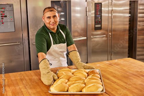 Portrait of hispanic smiling baker in a commercial kitchen of grocery placing tray of freshly baked bolillos from oven on to large worktable. photo