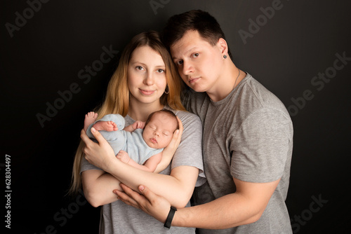 A cute newborn baby boy sleeps in a blue overalls in the first days of life. In the arms of father and mother. Warm parental hugs. On a black background. Professional portrait photography.