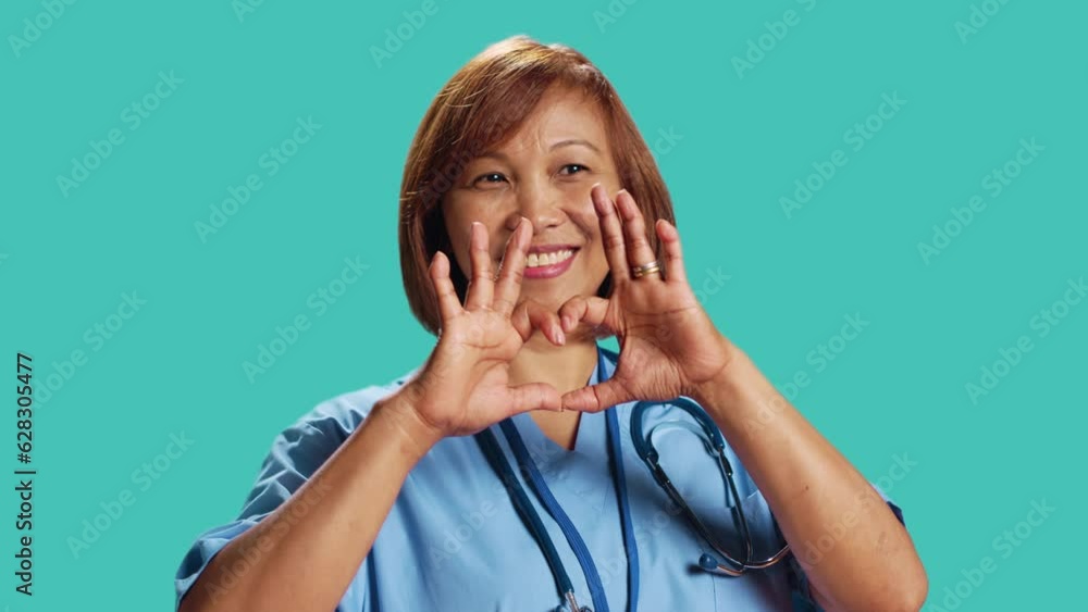 Close up shot of joyful qualified BIPOC nurse doing heart symbol shape with hands. Clinic employee showing love gesturing while at work, isolated over blue studio background Close up shot of joyful qualified BIPOC nurse doing heart symbol shape with hands. Clinic employee showing love gesturing while at work, isolated over blue studio background