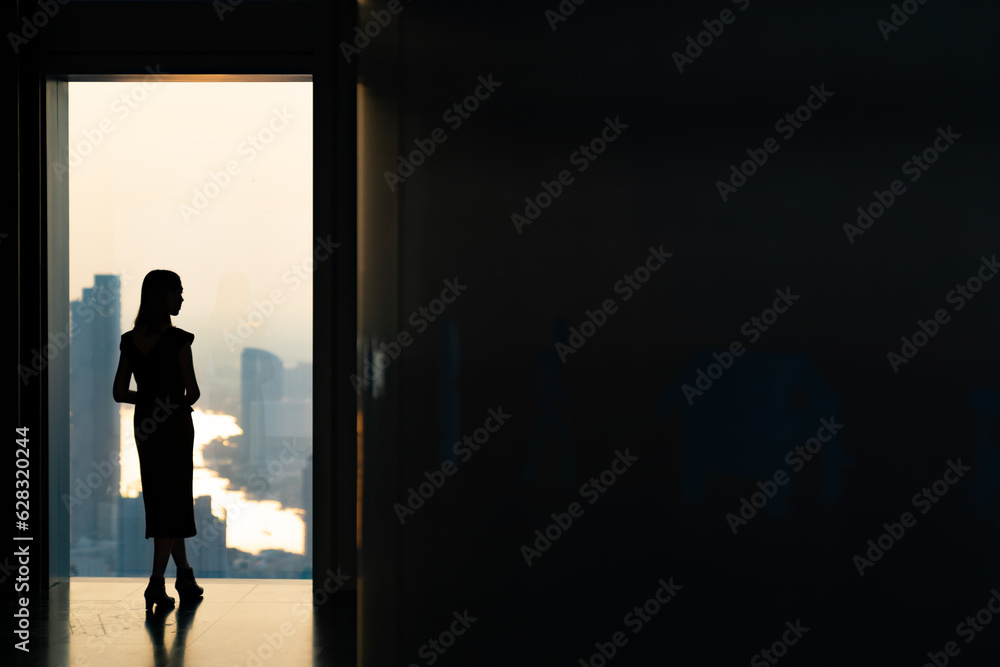 Fototapeta premium Silhouette Asian woman standing by the window at skyscraper building in metropolis and looking cityscape at summer sunset. Beautiful girl enjoy urban lifestyle activity in the city on holiday vacation