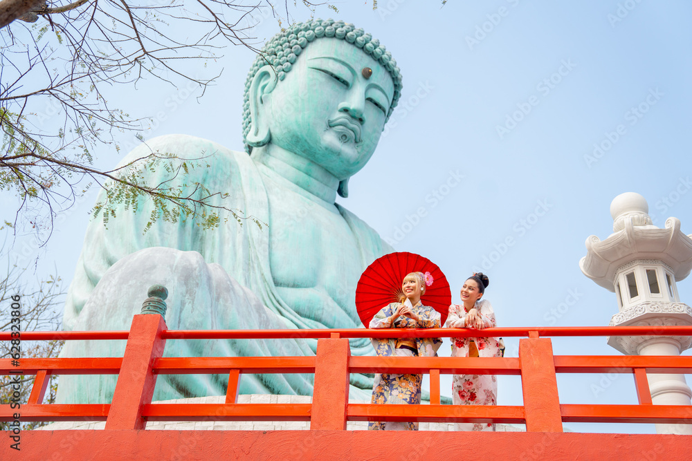 Fototapeta premium Wide shot of two Asian pretty women stay close to red railing in the area of base of green big buddha statue and they look to left side with happiness.