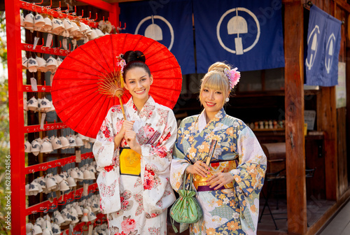 Close up two Asian women wear japanese style dress with one hold red umbrella and stand in front of small shop also look at camera with smiling.