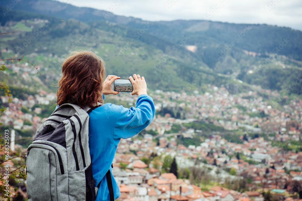 A hiker girl with a backpack inspects the sights of a beautiful European city in the mountains and takes photos on her smartphone A hiker girl with a backpack inspects the sights of a beautiful European city in the mountains and takes photos on her smartphone