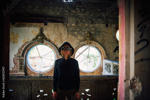 Valokuva A blonde girl in steampunk glasses and a black hoodie with a hood in an abandoned old building with unusual round windows