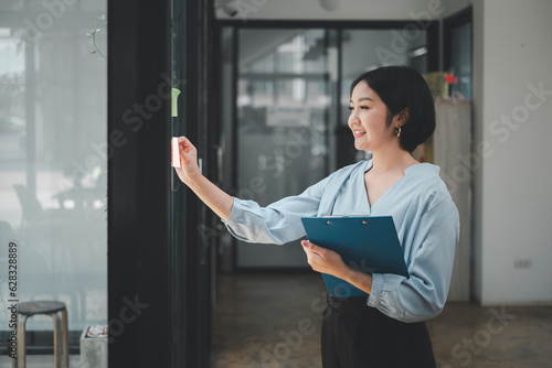 Foto Smiling businesswoman sticking adhesive notes on glass window in office