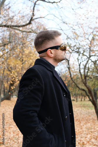 A man in a business suit, coat and steampunk goggles poses in an autumn park © ribalka yuli