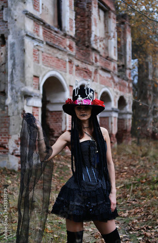 Female Halloween look. A woman in a black dress with a corset, a top hat decorated with skeleton figures poses for the camera against the backdrop of an old abandoned building
