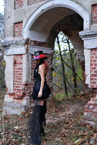 Female Halloween look. A woman in a black dress with a corset, a top hat decorated with skeleton figures poses for the camera against the backdrop of an old abandoned building