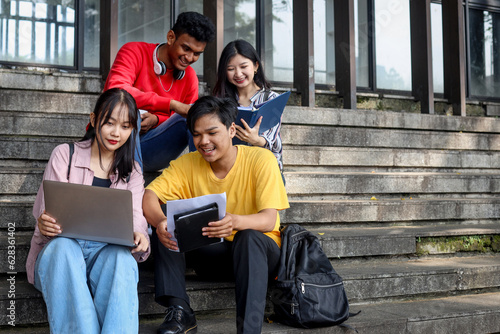 Young multiethnic college students reading, studying on laptop, preparing for exam or working on group project while sitting on staircase or steps of college campus photo