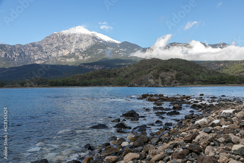 Deserted coast of the Gulf of the Sea. Curved coastline of dark sand and pebbles. Discarded snag on the shore. Mediterranean landscape, mountain ridge and Mount Olympus (aka Tahtali) in the background