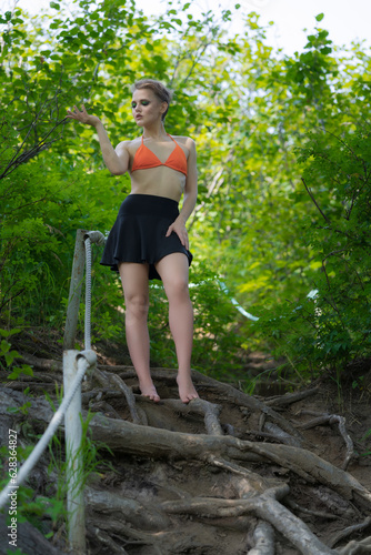 Caucasian woman standing and poses in forest on ladder made of tree roots. Sensuality adult female dressed in orange bikini top and mini skirt. Blonde model with short hair and bright make-up © Alexander Piragis