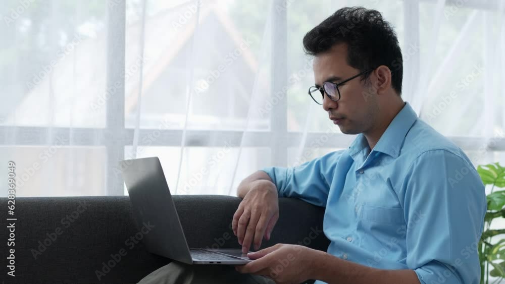 Asian business man using laptop, reading, researching, sitting on sofa at home, shopping or chatting online in social network, having fun, watching movies, freelancer working on computer project. Asian business man using laptop, reading, researching, sitting on sofa at home, shopping or chatting online in social network, having fun, watching movies, freelancer working on computer project.