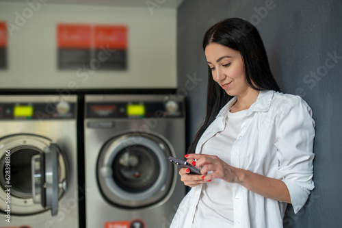 Fotografie Woman waiting in a laundry room using mobile phone, browsing internet