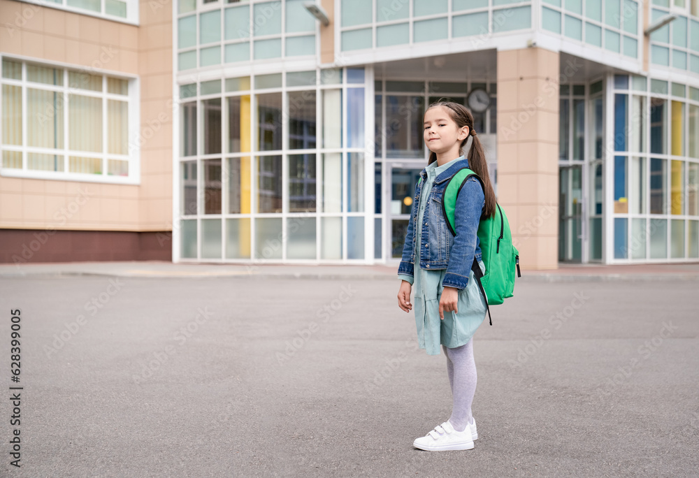 Little asian girl stands outside the school with a backpack on her back Little asian girl stands outside the school with a backpack on her back