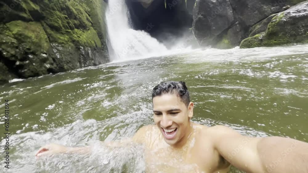 A young vlogger recording himselft while happily swimming into the lake near a waterfall, inside a rainforest. A young vlogger recording himselft while happily swimming into the lake near a waterfall, inside a rainforest.