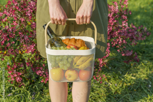 Faceless photo. Close-up of a girl's hands, preparing for a picnic, holding a white iron basket filled with fruits. It embodies the concept of abundance, harvest, nature care, and plastic-free living