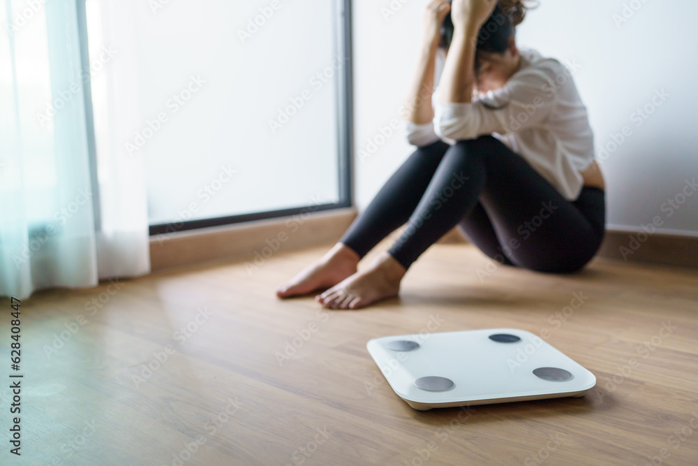 Fototapeta premium Sad woman with fat upset bored of dieting Weight loss fail Fat diet and scale sad asian woman on weight scale at home weight control