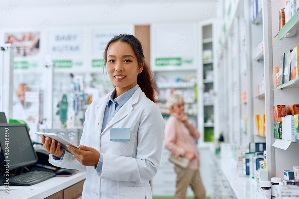 Female Chinese pharmacist using touchpad while working in drugstore and looking at camera. Female Chinese pharmacist using touchpad while working in drugstore and looking at camera.
