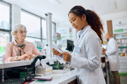 Young Chinese pharmacist reads prescription while serving senior woman in pharmacy.