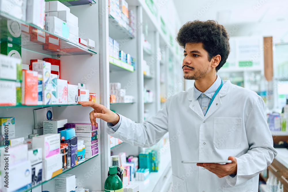 Young pharmacist goes through inventory of medicines on shelves in drugstore.