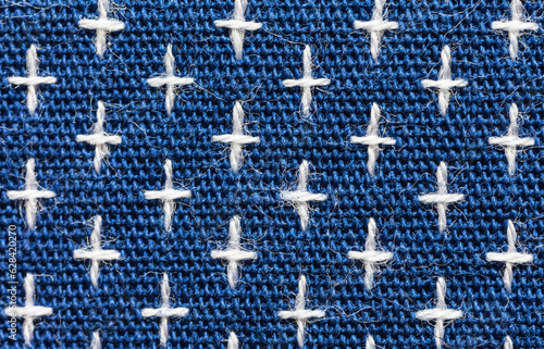 Close-up of machine embroidery sashiko with white crosses on blue background. Textile backdrop made of weaving natural cotton threads. Basis for sewing comfortable clothes, shirts, blouses. Macro photo