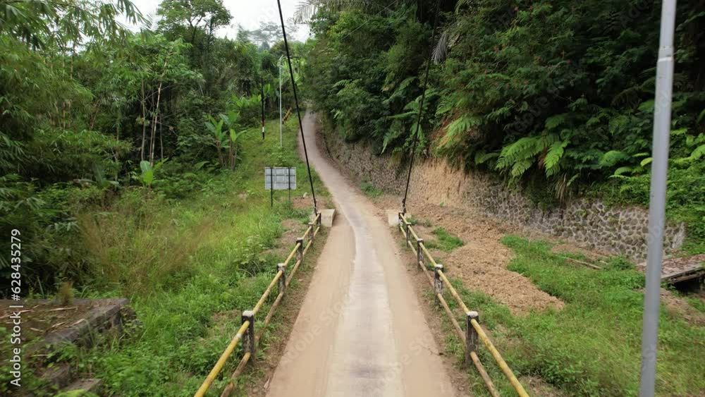 Aerial camera fly back along small suspension bridge, spanning across tropical ravine. Lush green thickets around, narrow roadway is for pedestrians and motorcycles only. Aerial camera fly back along small suspension bridge, spanning across tropical ravine. Lush green thickets around, narrow roadway is for pedestrians and motorcycles only.
