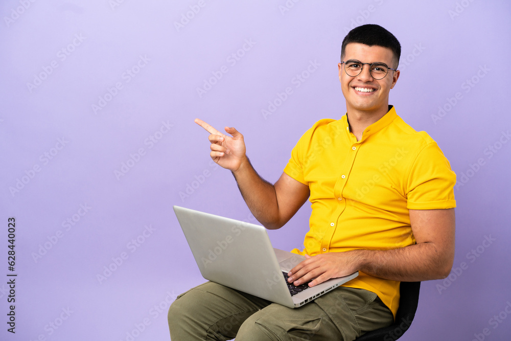 Young man sitting on a chair with laptop pointing finger to the side Young man sitting on a chair with laptop pointing finger to the side