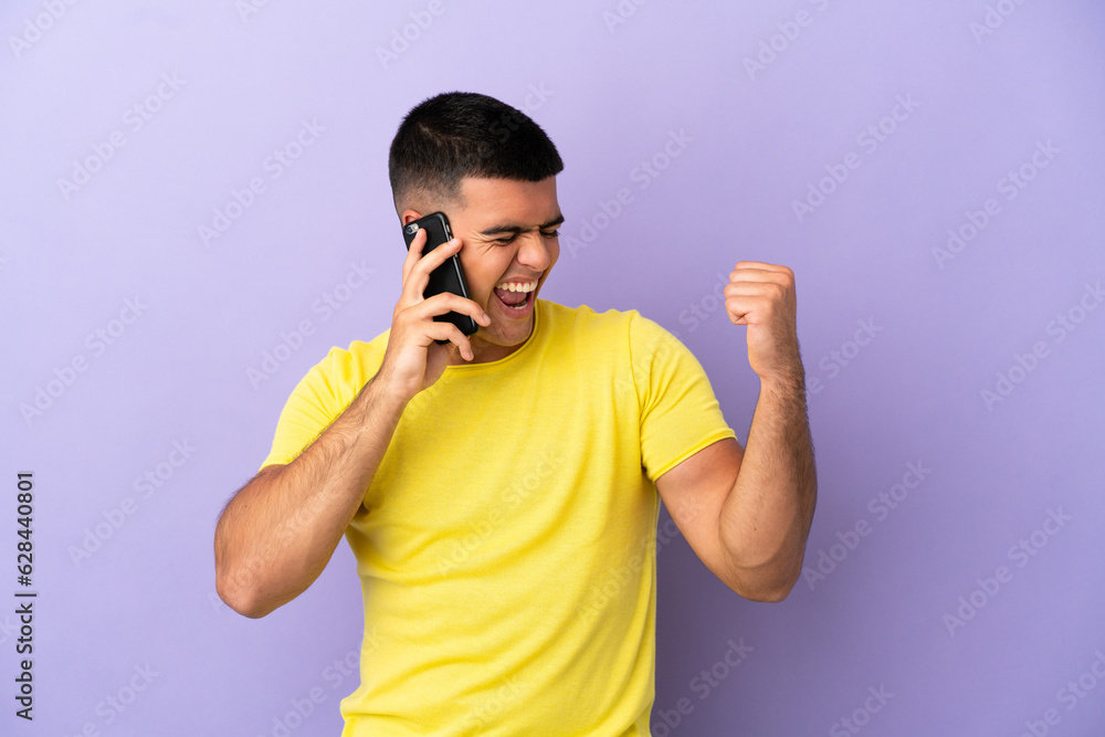 Young handsome man using mobile phone over isolated purple background celebrating a victory Young handsome man using mobile phone over isolated purple background celebrating a victory