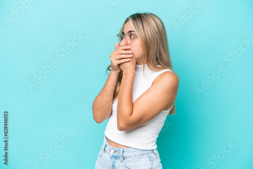 Young caucasian woman isolated on blue background covering mouth and looking to the side