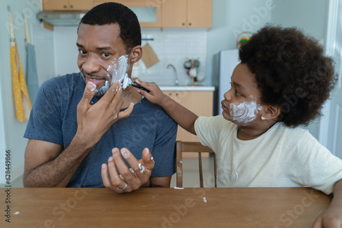 Happy African American Father and little son having fun while shaving and looking at mirror with kid holding razor helping to shave on his father at home. Black Family Activity together.