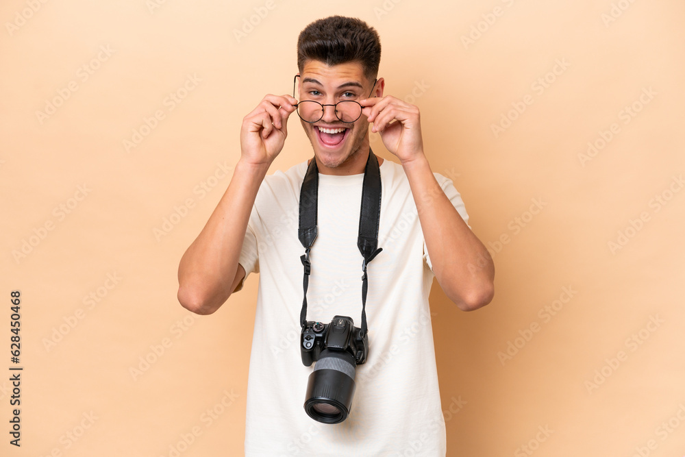 Young photographer caucasian man isolated on beige background with glasses and surprised Young photographer caucasian man isolated on beige background with glasses and surprised