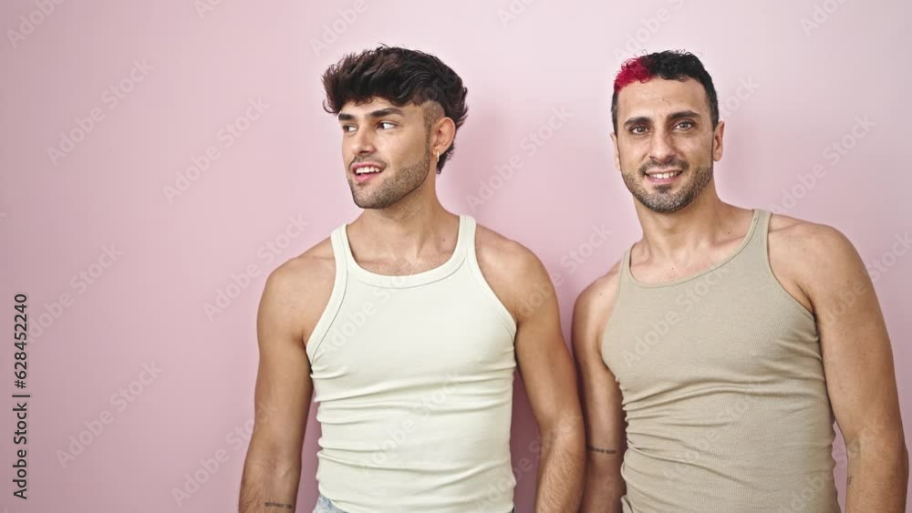 Two men couple standing with surprise expression pointing to side over isolated pink background Two men couple standing with surprise expression pointing to side over isolated pink background