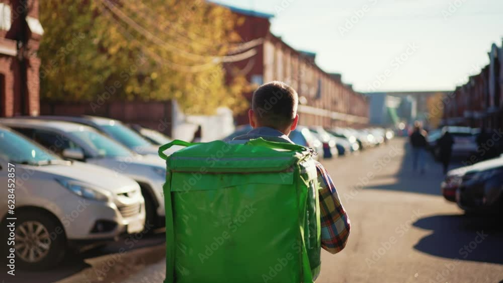 Man courier finding address on map in smartphone on city street looking around. Food delivery guy navigates in phone application with green huge backpack on spine. Fast delivery, courier work concept. Man courier finding address on map in smartphone on city street looking around. Food delivery guy navigates in phone application with green huge backpack on spine. Fast delivery, courier work concept.