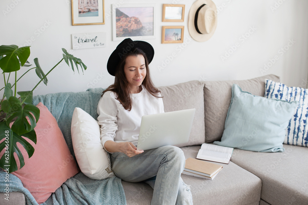 Young woman using laptop, working online, wearing black hat, smiling, sitting on a comfy couch. Trendy hipster cozy interior. Artist living slow freelance life. Millennial lifestyle. Young woman using laptop, working online, wearing black hat, smiling, sitting on a comfy couch. Trendy hipster cozy interior. Artist living slow freelance life. Millennial lifestyle.