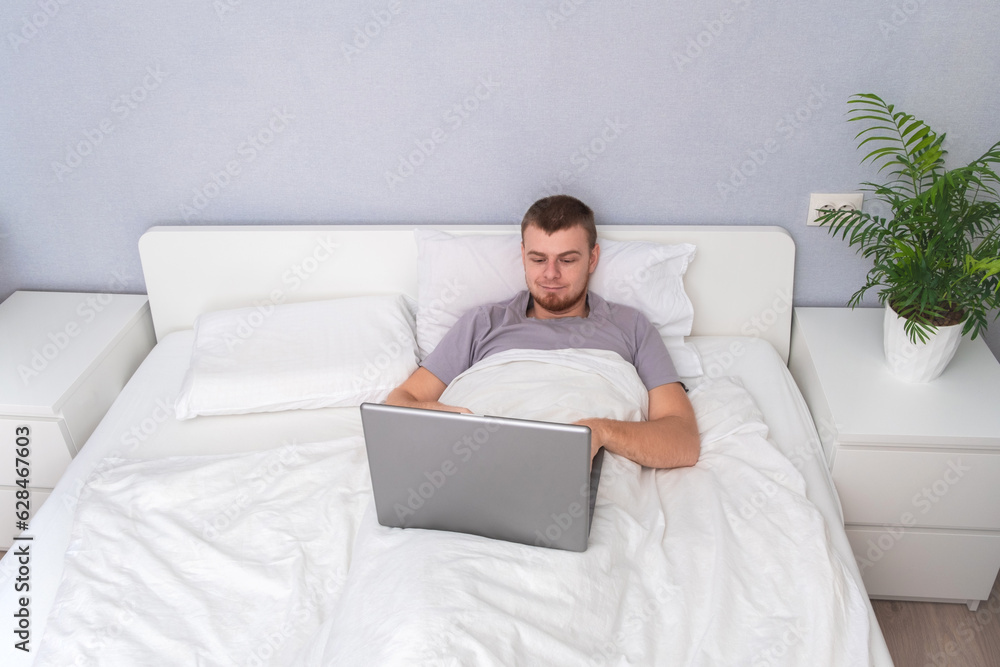 A man with laptop is lying in bed. Freelancer works remotely from home. Distance education. Online shopping without leaving home. Internet surfing. Gambling addiction. Black Friday sale. Cyber Monday. A man with laptop is lying in bed. Freelancer works remotely from home. Distance education. Online shopping without leaving home. Internet surfing. Gambling addiction. Black Friday sale. Cyber Monday.