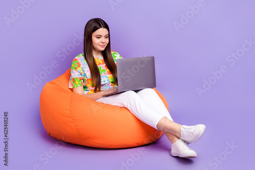 Full body length photo of satisfied young girl concentrated sit cozy bean bag netbook chatting online isolated on violet color background
